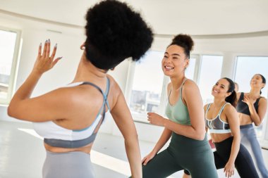 Spor kıyafetli bir grup genç kadın, kadın eğitmenle pilates yapıyor, el sallıyor.
