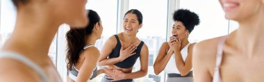 focus on diverse female friends in sportswear laughing while chatting before pilates class, banner clipart