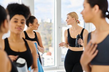 Mutlu sarışın kadın spor şişesi tutuyor ve yoga stüdyosundaki Afrikalı Amerikalı arkadaşıyla konuşuyor.