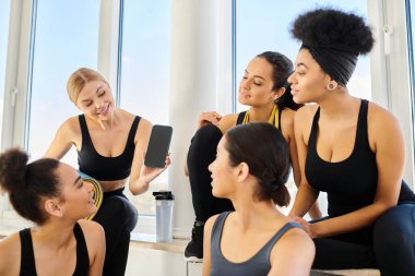 Genç sarışın kadın yoga stüdyosunda idmandan sonra kız arkadaşlarına akıllı telefon gösteriyor.