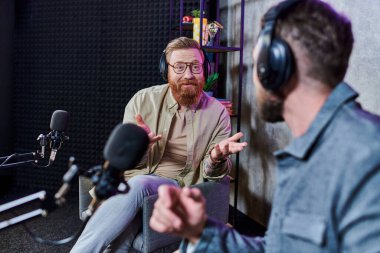 Sakallı yakışıklı mülakatçı ve kulaklıklı konuğu stüdyoda sorular, podcast hakkında konuşuyorlar.