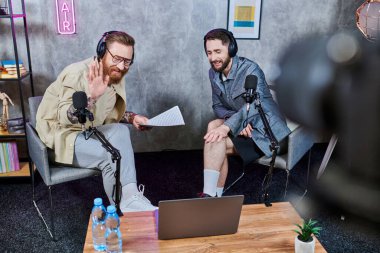 Podcast sırasında kulaklıklı, sıradan giyinmiş iki yakışıklı sakallı adam aktif olarak konuşuyor.
