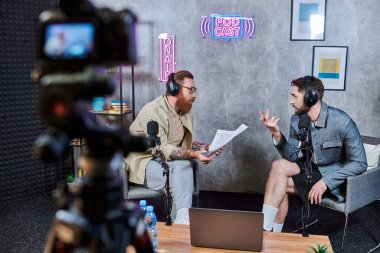 Podcast sırasında kulaklıklı, sıradan giyinmiş iki yakışıklı sakallı adam aktif olarak konuşuyor.