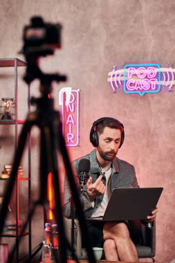 İyi görünümlü sakallı, rahat giyimli bir adam stüdyoda podcast sırasında dizüstü bilgisayarla oturuyor.
