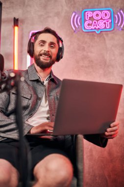 Her gün rahat giyinen yakışıklı sakallı adam podcast sırasında dizüstü bilgisayarla stüdyoda oturuyor.