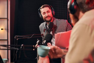 Elinde kahve fincanı olan neşeli sakallı adam podcaster 'ının yanındaki evraklara bakıyor.
