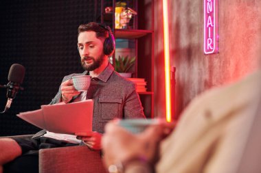 Elinde kahve fincanı olan yakışıklı sakallı adam podcaster 'ının yanındaki evraklara bakıyor.