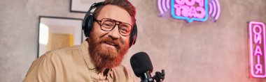 Kırmızı saçlı, kulaklıklı neşeli yakışıklı adam podcast sırasında kameraya bakıyor.