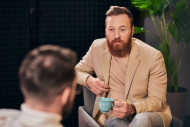 good looking red haired man with coffee cup sitting during discussion next to his interviewer clipart