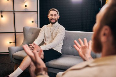 cheerful bearded men in casual outfit smiling and looking at his interviewer while sitting in studio clipart