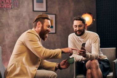 two cheerful elegant men with beards in chic clothes looking at smartphone during interview clipart