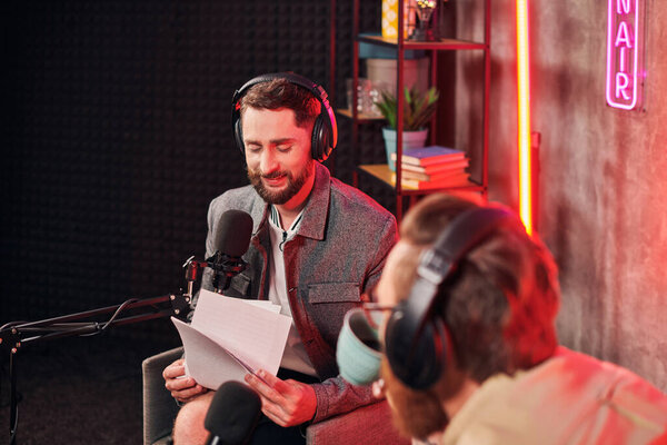 веселый бородатый мужчина с кофейной чашкой в руках, глядя на бумажную работу рядом со своим подкастером