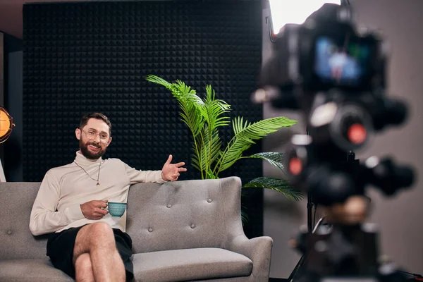 cheerful bearded man in elegant attire with glasses holding coffee cup during interview in studio