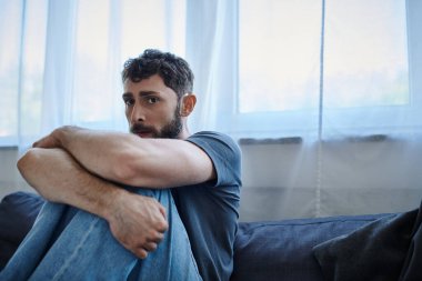 Ruhsal çöküntü sırasında kanepede oturup kameraya bakan sıradan giyinmiş sakallı, endişeli bir adam.