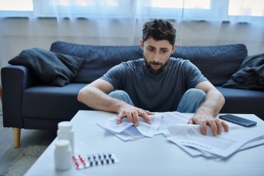 Depresyonda olan sakallı hasta bir adam depresif bir olay sırasında üzerinde kağıtlar ve haplarla masada oturuyor.