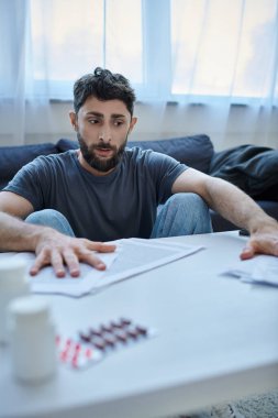 Depresyonda olan sakallı hasta bir adam depresif bir olay sırasında üzerinde kağıtlar ve haplarla masada oturuyor.