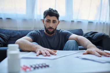 Depresyonda olan sakallı hasta bir adam depresif bir olay sırasında üzerinde kağıtlar ve haplarla masada oturuyor.