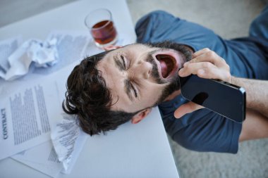 Sinir krizi sırasında yanında bir bardak alkolle akıllı telefona bağıran sinirli bir adam.