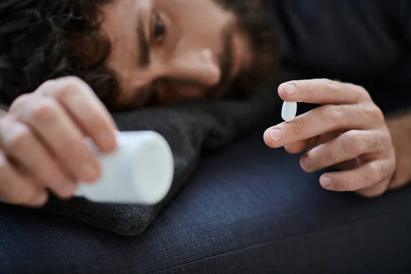 ill suffering man in casual attire lying on sofa with pills in hand, mental health awareness