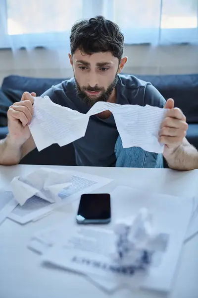 frustrated man with beard sitting at table with papers and phone on it during mental breakdown