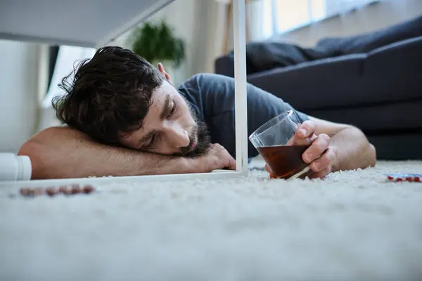 stock image depressed man in casual attire drinking alcohol and looking at pills during mental breakdown