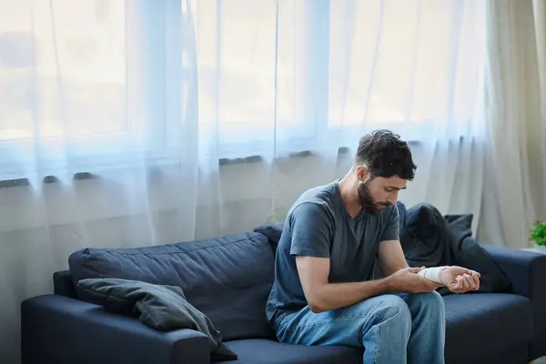 Stock image depressed man with bandage on arm after attempting suicide sitting on sofa, mental health awareness