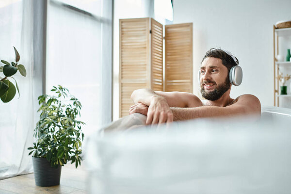 joyful attractive man with beard and headphones sitting and relaxing in his bathtub, mental health