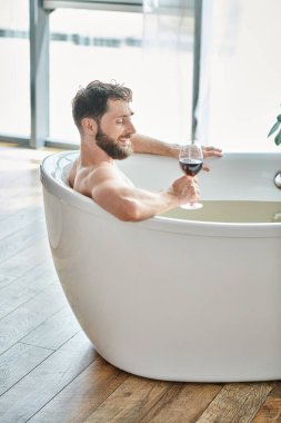 Banyo küvetinde bir bardak kırmızı şarapla rahatlayan neşeli, yakışıklı adam, zihinsel sağlık farkındalığı.