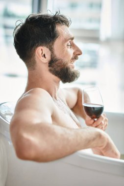 Mutlu, sakallı, küvette bir bardak kırmızı şarapla rahatlayan, zihinsel sağlığın farkında olan çekici bir adam.