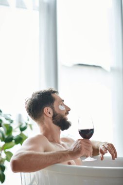 Sakallı ve göz bantlı neşeli yakışıklı adam elinde kırmızı şarapla küvette dinleniyor.