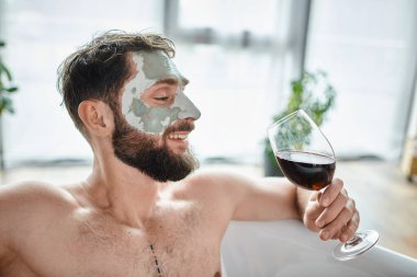 Sakallı ve yüz maskeli neşeli çekici bir adam kırmızı şarapla küvette dinleniyor.
