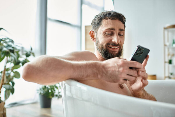 merry handsome man with beard looking at his smartphone while in bathtub, mental health awareness