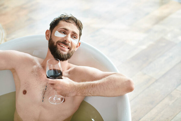 joyful attractive man with beard and eye patches relaxing in bathtub with glass of red wine