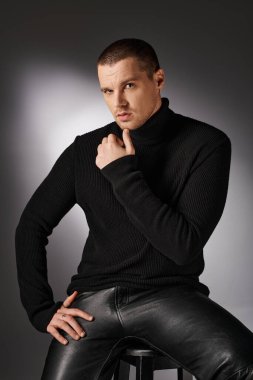 pensive handsome stylish man in black turtleneck looking at camera on grey backdrop with lighting clipart