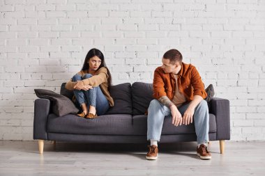 frustrated interracial couple looking at each other on couch in living room, divorce concept clipart