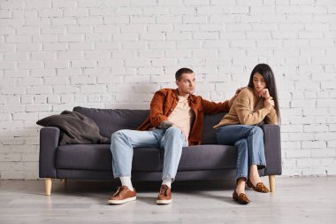 young man calming upset asian wife sitting on couch in living room, relationship difficulties clipart