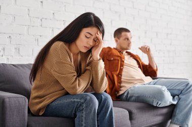 young depressed asian woman sitting with closed eyes near offended husband on couch in living room clipart