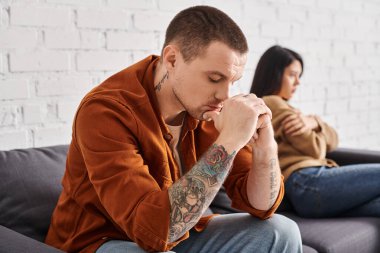 Depresyondaki dövmeli adam. Gözleri kapalı. Evdeki koltukta oturan dargın Asyalı eş, boşanma kavramı.