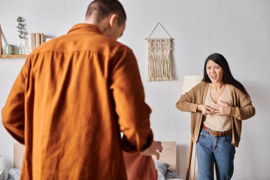 irritated asian woman quarreling at husband standing with clothes in bedroom, family conflict clipart