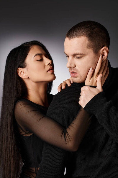 чувственная азиатская брюнетка в черном модном одеянии обнимающая красивого мужчину на сером фоне