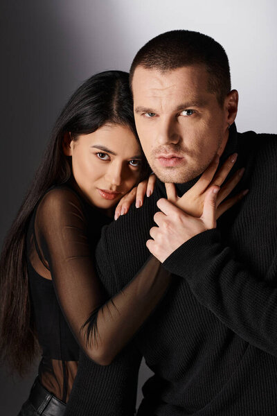 романтическая межрасовая пара в черном модном одеянии обнимающая и смотрящая в камеру на сером