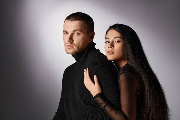 young trendy interracial couple in black outfit looking at camera on grey backdrop with lighting