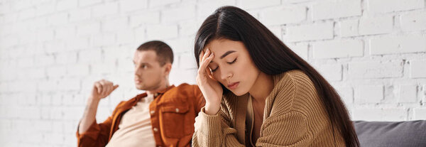 расстроен азиатская женщина сидит с закрытыми глазами рядом мрачный муж на диване в гостиной, баннер