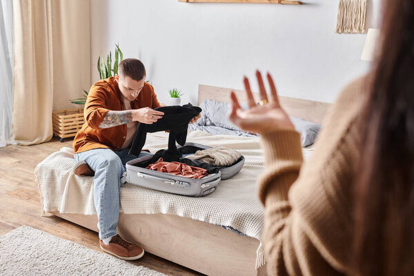 offended tattooed man packing suitcase near wife quarreling in bedroom at home, misunderstanding