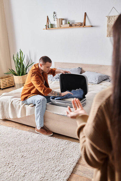 gloomy tattooed man packing suitcase near wife quarreling in bedroom at home, misunderstanding