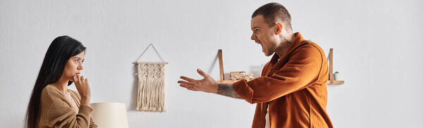 young and irritated man quarreling at asian wife near suitcase and belongings in bedroom, banner