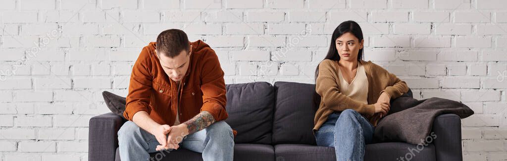 Young offended interracial couple sitting on couch in living room, family divorce concept, banner