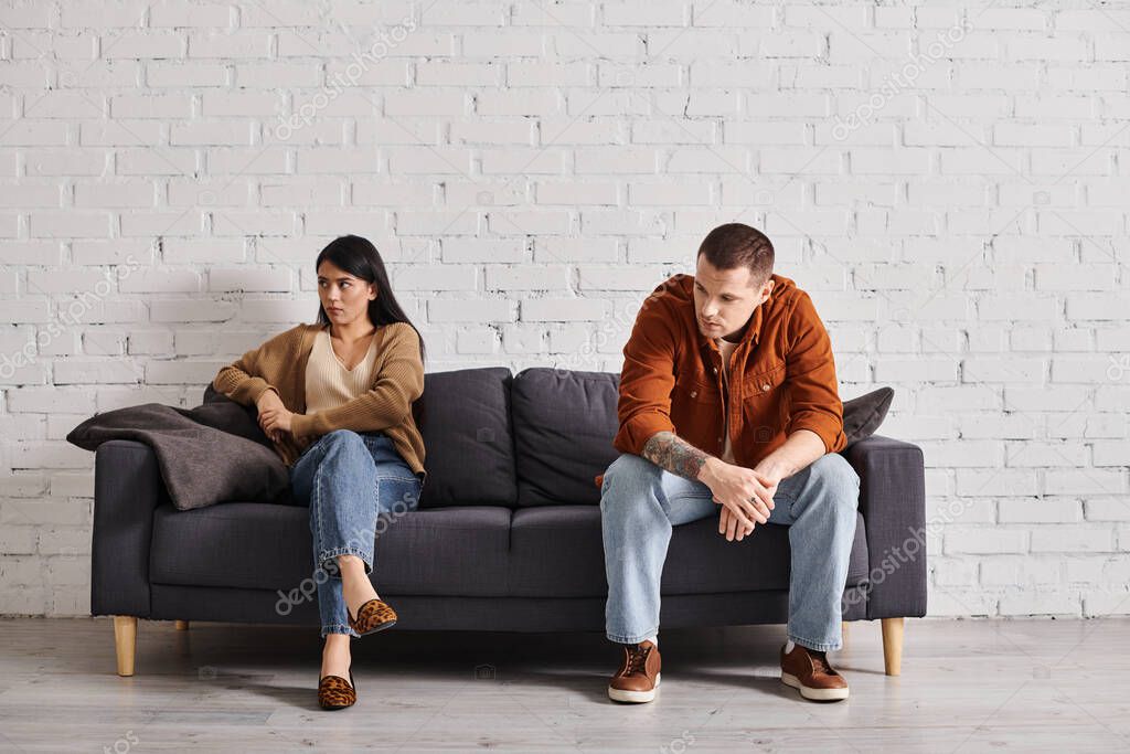 Young upset interracial couple sitting on couch in living room at home, family divorce concept