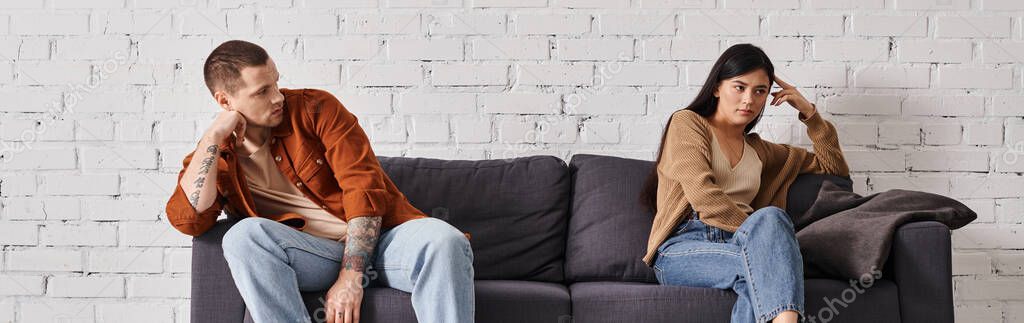 Sad multicultural couple sitting on couch in living room at home, family divorce concept, banner