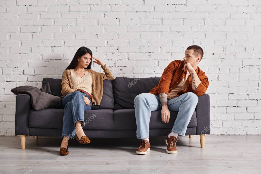 Offended interracial couple looking at each other on couch in living room, relationship difficulties